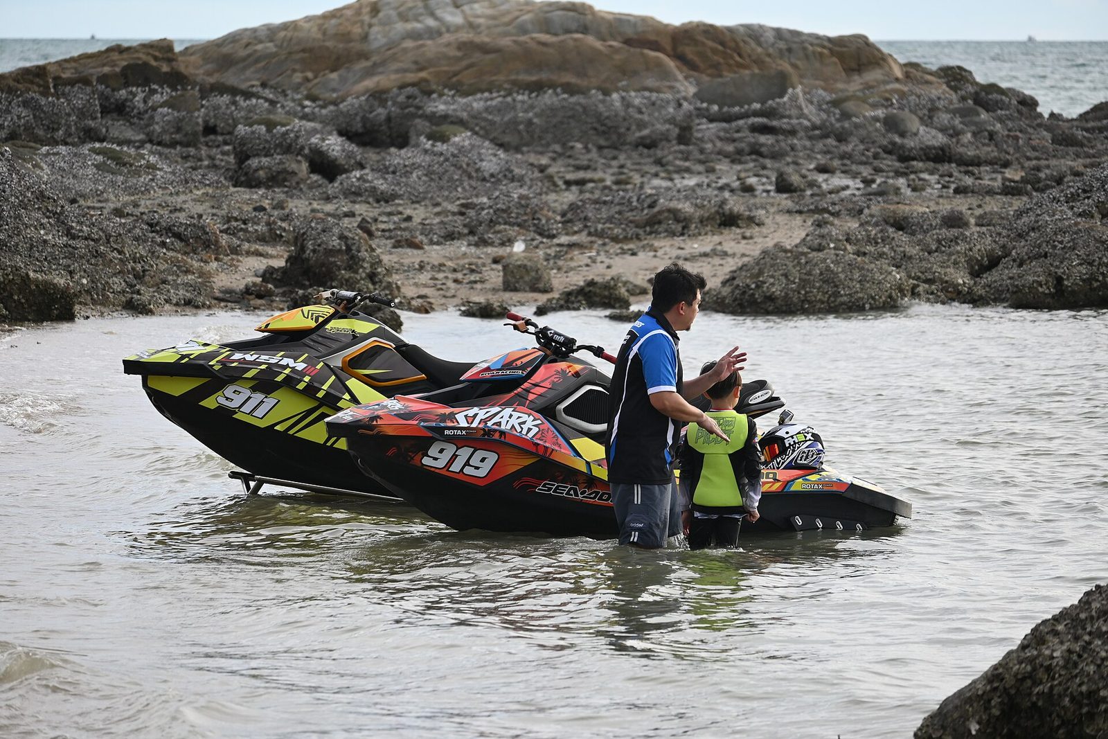 DFC 0386 A man helps a child board a colorful jet ski in shallow coastal waters near rocky outcrops.jpg
